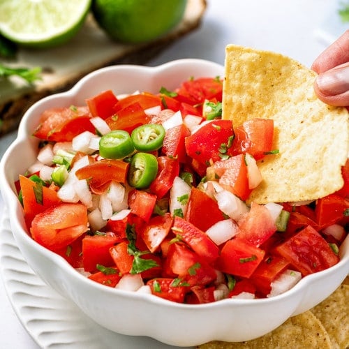 Hand scooping fresh pico de gallo with a tortilla chip from a bowl garnished with lime and green chiles.