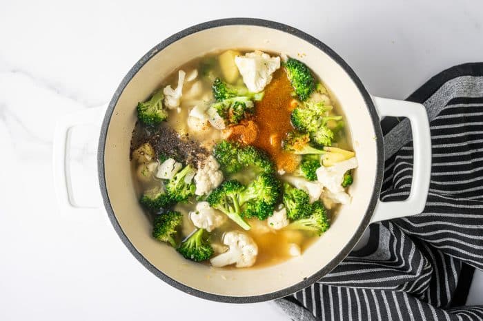Overhead shot of a cream enameled Dutch oven filled with broth covering broccoli, cauliflower, and potato pieces. A mound of ground black pepper and a pile of reddish-orange spice (cayenne) sit on top of the liquid near the surface. Black-and-white striped towel rests along the right edge.