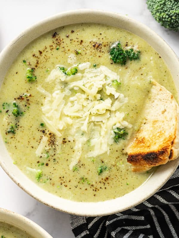 Overhead photo of a light beige bowl filled with creamy pale green broccoli cauliflower soup topped with shredded cheddar and cracked black pepper. A toasted bread wedge is tucked into the right side of the bowl for dipping. A second bowl is partially visible at the bottom left, with a black-and-white striped towel underneath and toast slices in the background.
