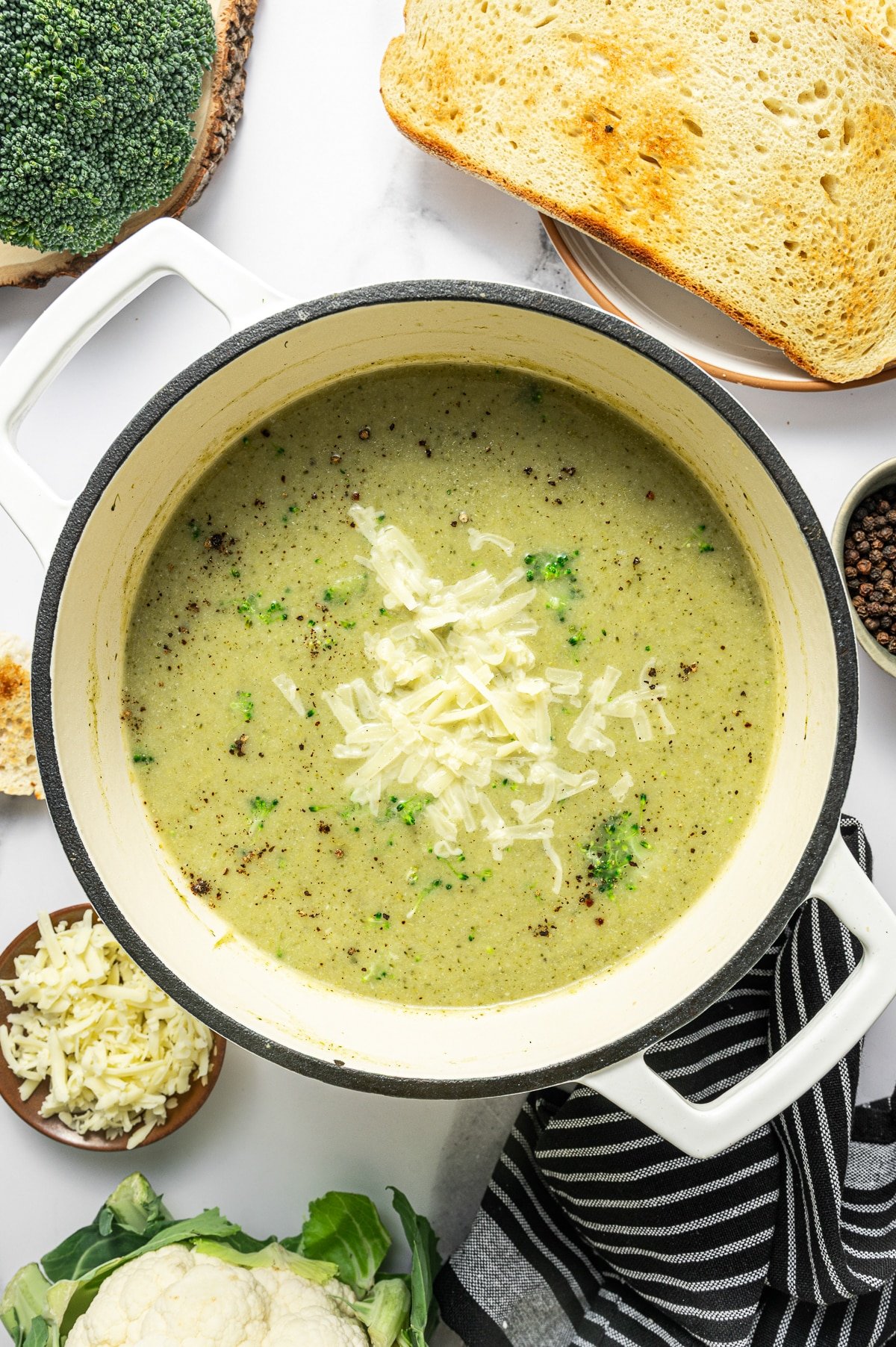 Overhead hero photo of broccoli cauliflower soup in a cream enameled Dutch oven. The soup is creamy pale green and topped with shredded cheddar, scattered black pepper, and a few small broccoli florets. Styled props surround the pot including toasted bread slices on a plate at the top right, a small bowl of shredded cheese at the bottom left, a small bowl of whole peppercorns on the right, and broccoli and cauliflower heads near the edges. A black-and-white striped towel is draped along the lower right.
