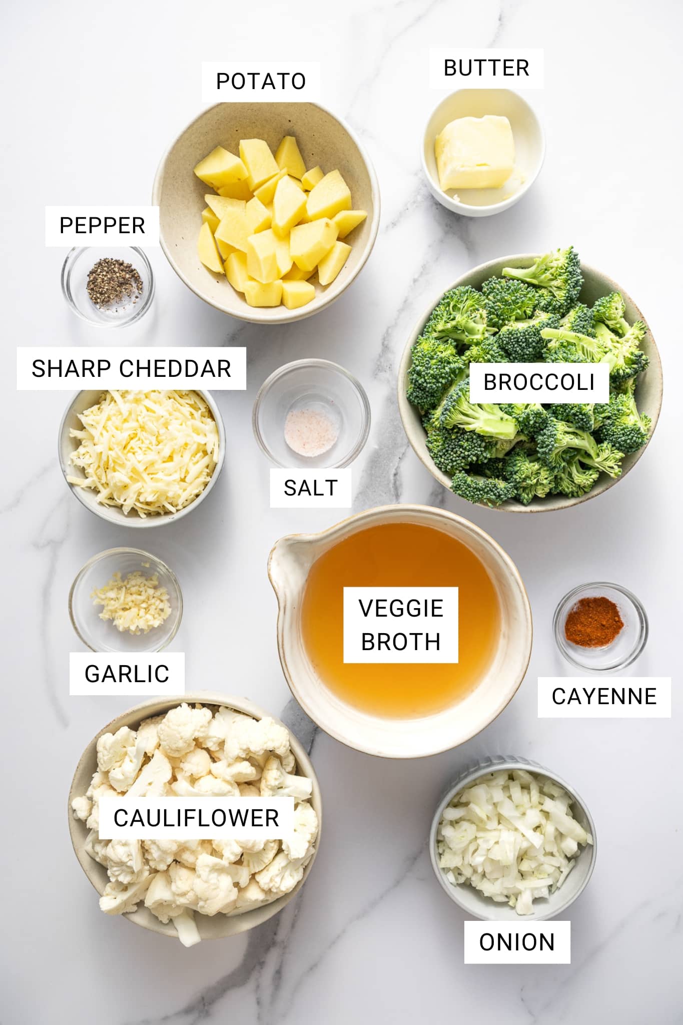 Overhead ingredient photo on a white marble surface with text overlays labeling each item: POTATO, BUTTER, PEPPER, SHARP CHEDDAR, SALT, BROCCOLI, VEGGIE BROTH, CAYENNE, GARLIC, CAULIFLOWER, and ONION. Ingredients are arranged in small bowls, including diced peeled potatoes in a beige bowl, broccoli florets in a beige bowl, cauliflower florets in a beige bowl, chopped onion in a white bowl, shredded sharp cheddar in a small bowl, minced garlic in a small glass bowl, salt and black pepper in small glass bowls, cayenne in a small glass bowl, and veggie broth in a cream-colored measuring jug.