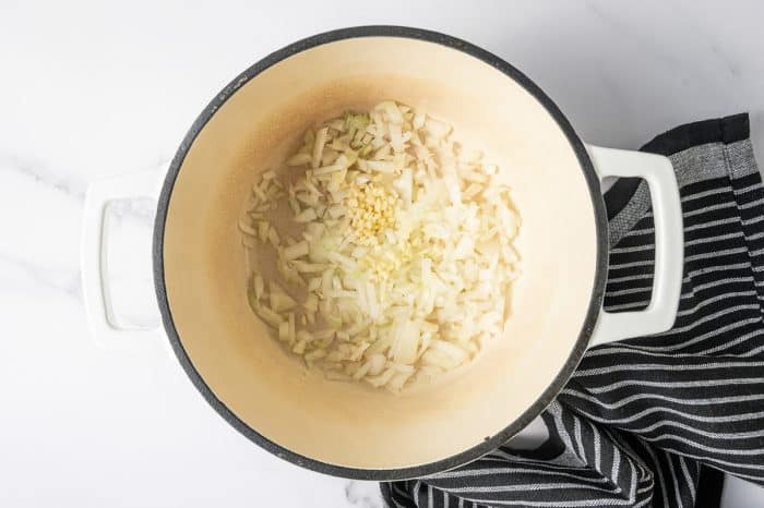 Overhead shot of a cream-colored enameled Dutch oven with a black rim and white handles. Chopped onion and minced garlic are sautéing in the bottom of the pot. The pot sits on a bright white background with a black-and-white striped kitchen towel tucked along the right side.