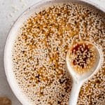 A top-down view of sesame dressing with a spoon resting in the bowl, featuring a heavy scattering of sesame seeds on the surface and styled with ginger root on the side. Bold text overlay highlights the recipe name.