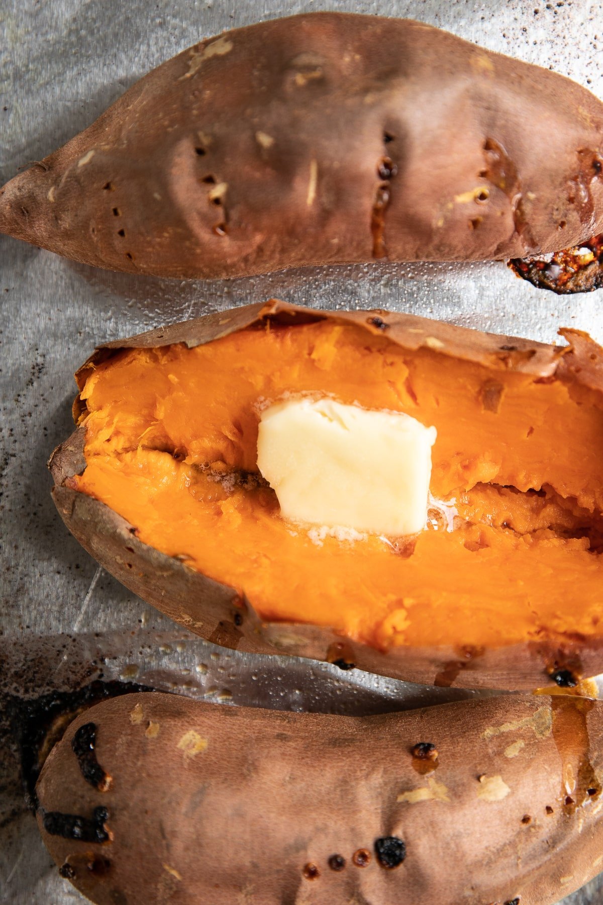Baked sweet potatoes on a baking sheet one is sliced open and garnished with butter.