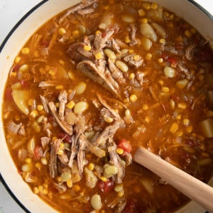 Large Dutch oven filled with a stew made with shredded pork, frozen lima beans, corn, tomatoes, potatoes, onions, and bbq sauce.