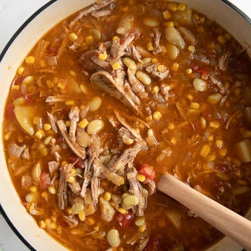 Large Dutch oven filled with a stew made with shredded pork, frozen lima beans, corn, tomatoes, potatoes, onions, and bbq sauce.