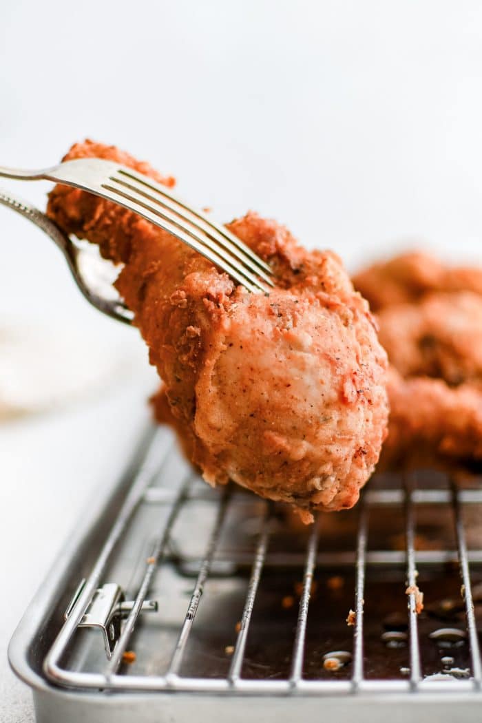 Two forks removing one crispy fried buttermilk chicken drumstick from a wire cooling rack.