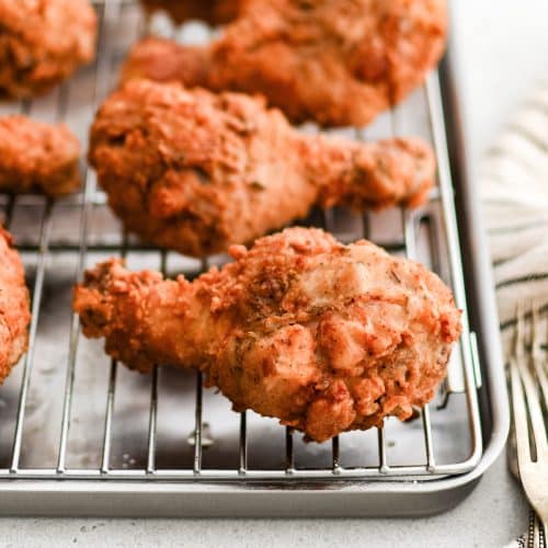 Crispy, golden buttermilk fried chicken drumsticks are cooling on a wire rack on a baking sheet.