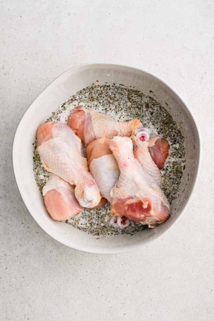 Raw chicken drumsticks in a large white bowl filled with seasoned buttermilk marinade.