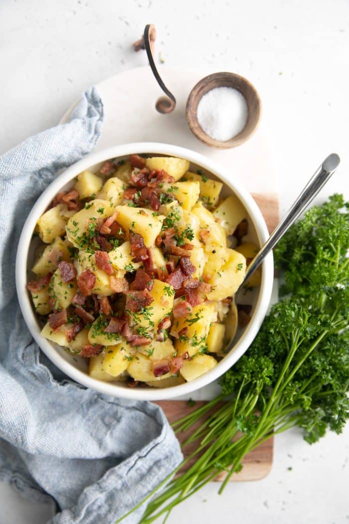 White serving bowl filled with German potato salad and garnished with extra bacon pieces and chopped parsley.