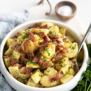 White serving bowl filled with German potato salad and garnished with extra bacon pieces and chopped parsley.