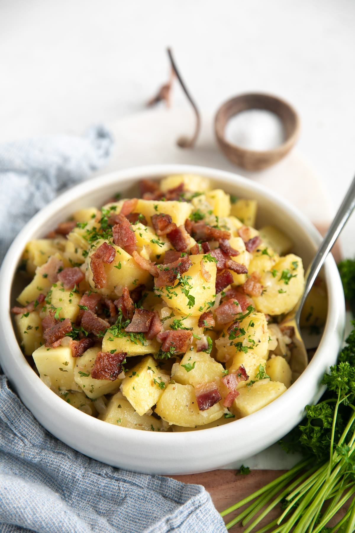 White serving bowl filled with German potato salad and garnished with extra bacon pieces and chopped parsley.