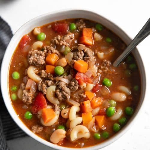 White serving bowl filled with filled with Hamburger Soup made with ground beef, tomatoes, macaroni noodles, peas, and carrots.