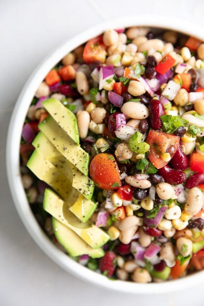 Bean salad filled with several types of beans, tomatoes, red onion, and sliced avocado.