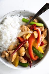 Szechuan chicken with colored bell peppers served with white rice in a white bowl.