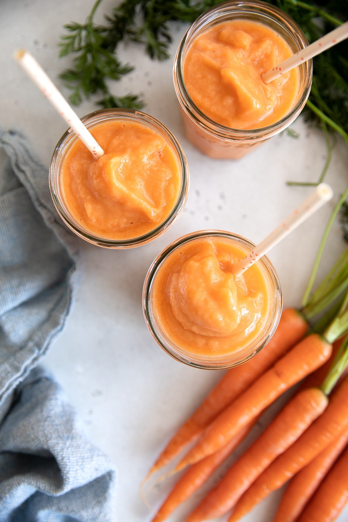 Overhead image of three glasses filled with icy cold carrot smoothie made with carrot juice, ginger, mango, and pineapple.