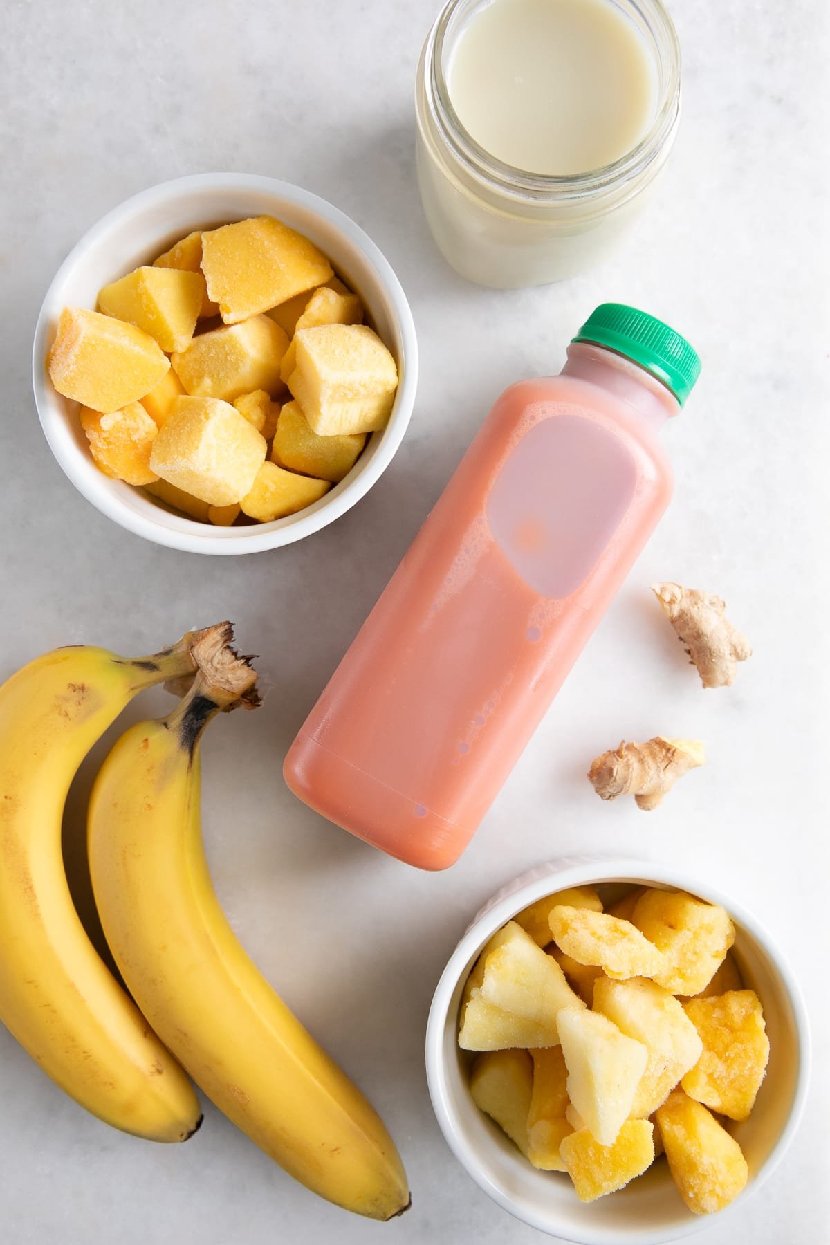Ingredients needed to make a carrot smoothie including frozen mango, pineapple, banana, carrot juice, ginger, and oat milk.