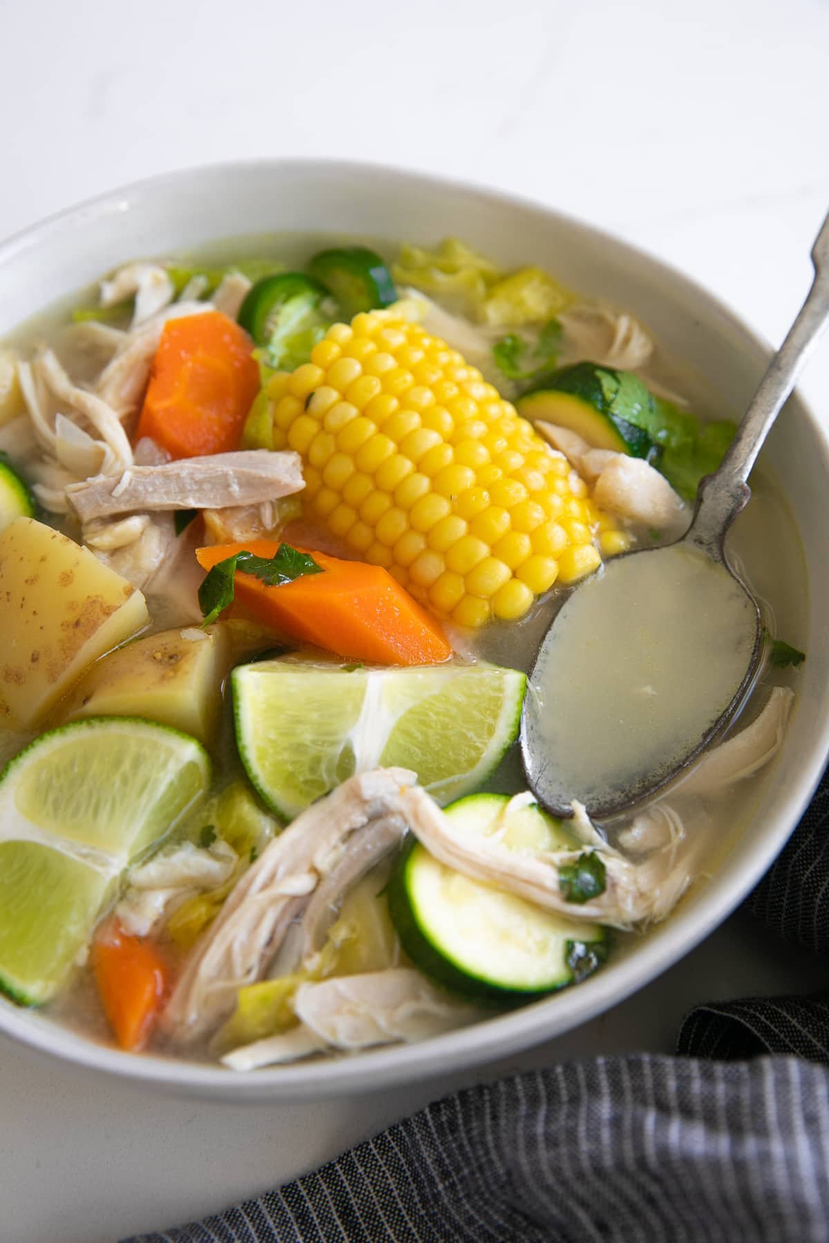 Large white shallow bowl filled with caldo de pollo made with chicken, carrots, corn, lime juice, potatoes, and zucchini.
