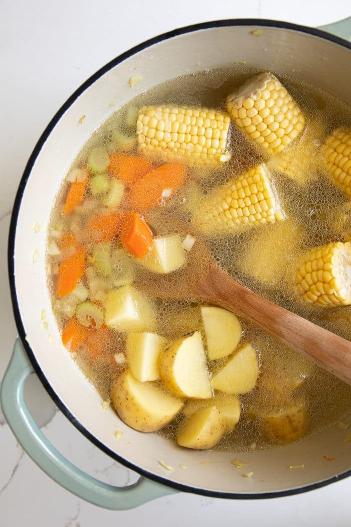 Diced potatoes and halved corn on the cob with carrots, celery, onions, and garlic in a large Dutch oven filled with chicken broth.