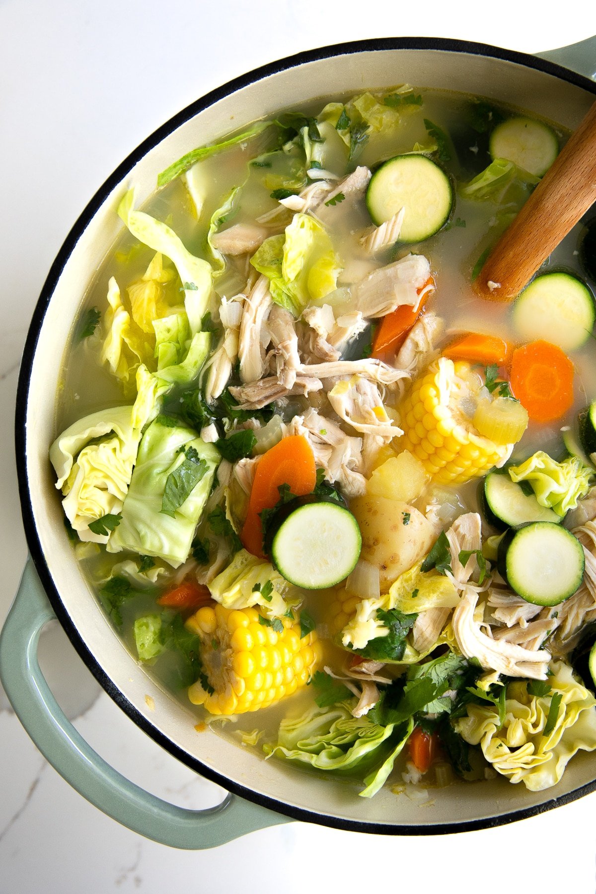 Large pot of soup filled with a clear chicken broth, zucchini, carrots, corn, shredded chicken, cabbage and potatoes.