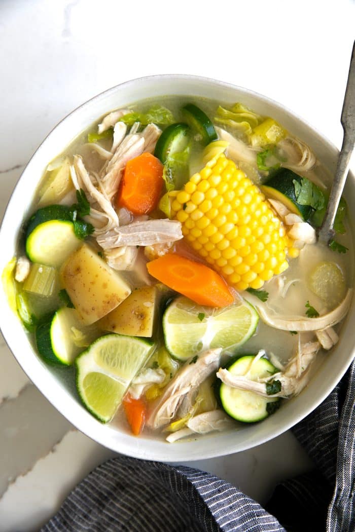 Large white shallow bowl filled with caldo de pollo made with chicken, carrots, corn, lime juice, potatoes, and zucchini.