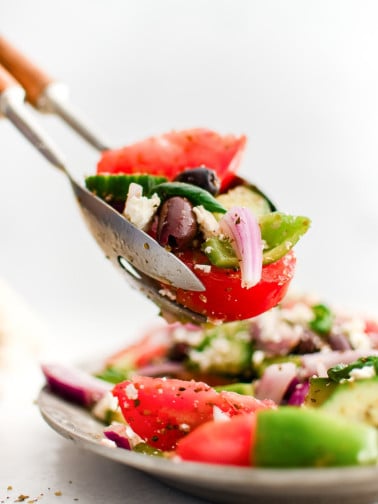 Salad tongs lifting a serving of Greek salad from the white platter.