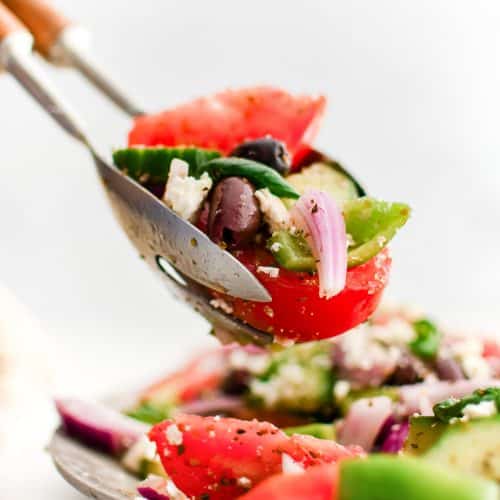 Salad tongs lifting a serving of Greek salad from the white platter.