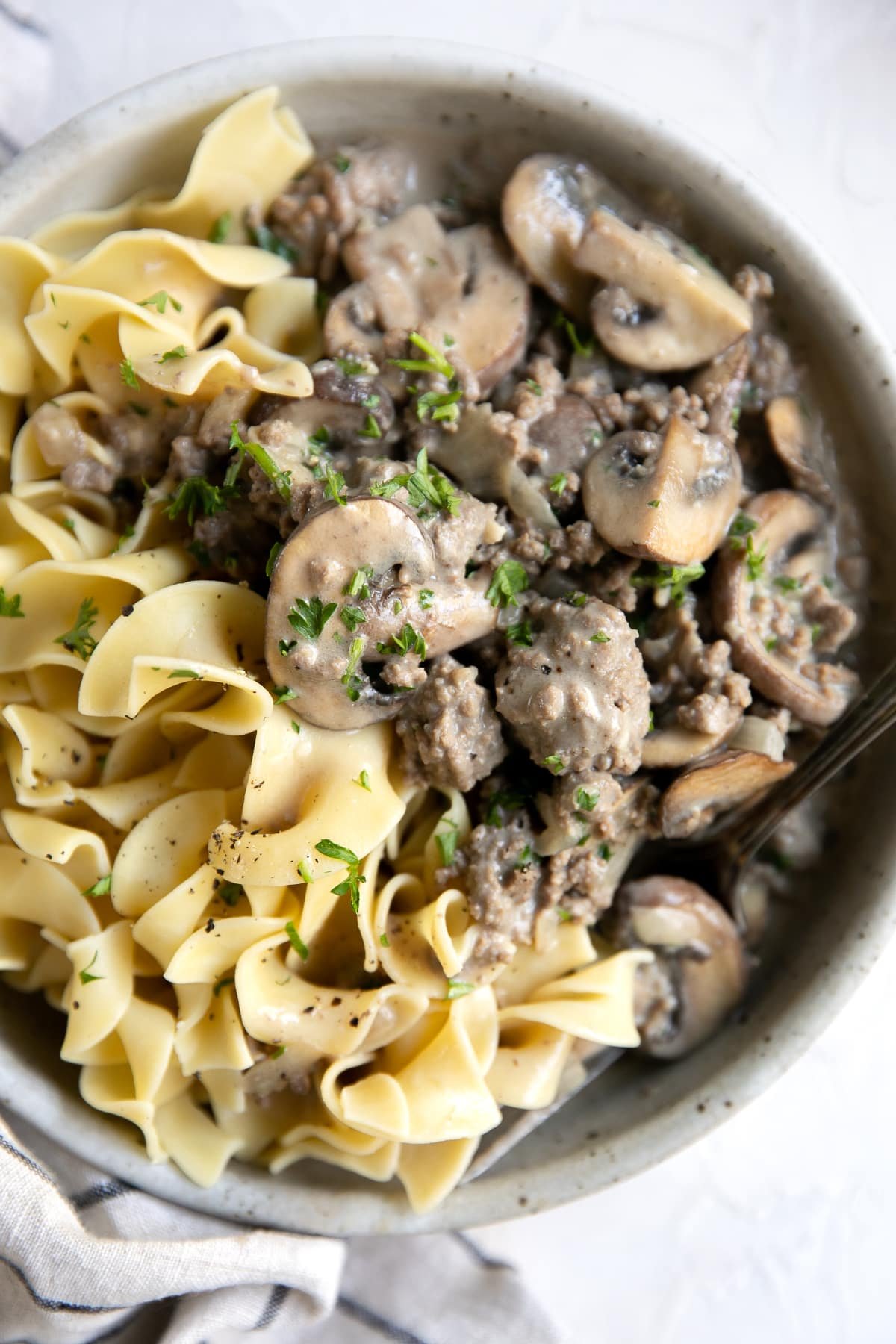 Large white serving dish filled with egg noodles and hamburger stroganoff garnished with fresh parsley.