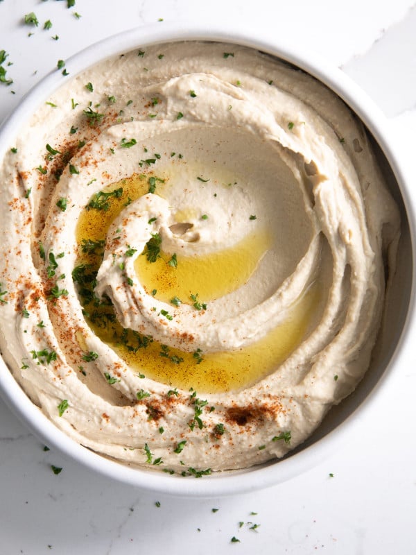 White bowl filled with the best creamy classic hummus and garnished with olive oil, paprika, and minced parsley.