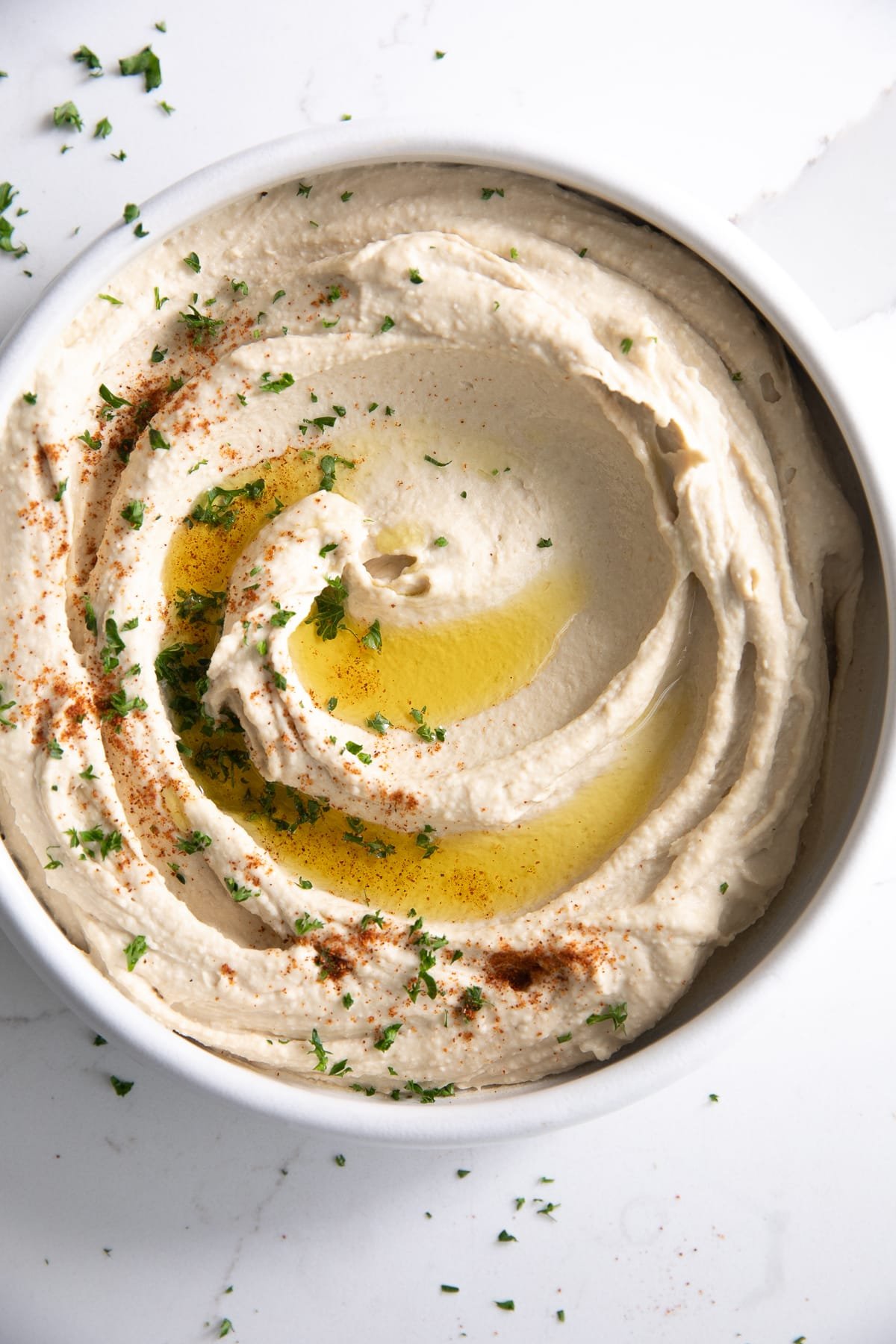 White bowl filled with creamy classic hummus drizzled with olive oil and sprinkled with fresh parsley and paprika.