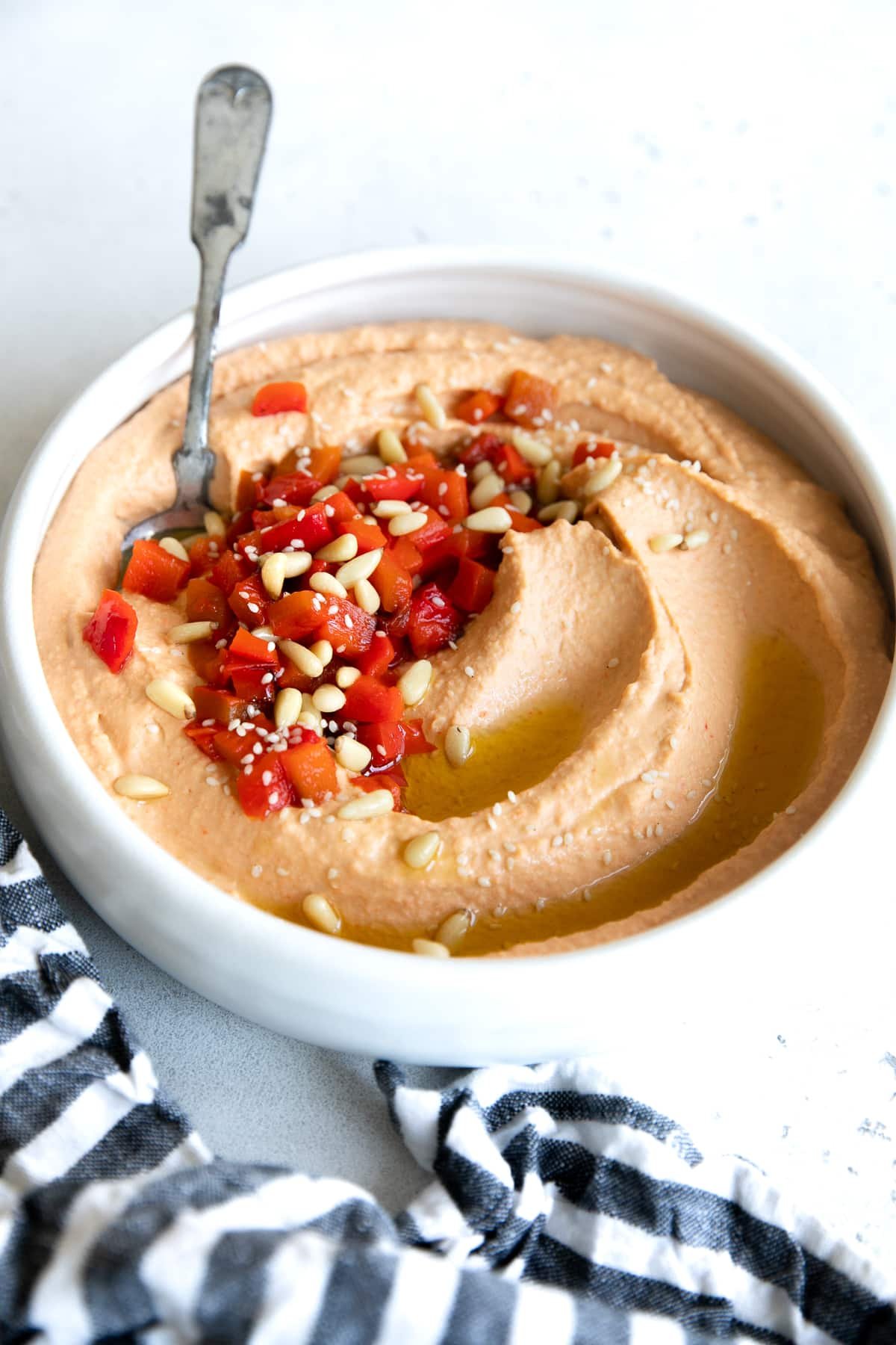 Roasted red pepper hummus in a white bowl topped with chopped roasted red bell pepper and pine nuts.