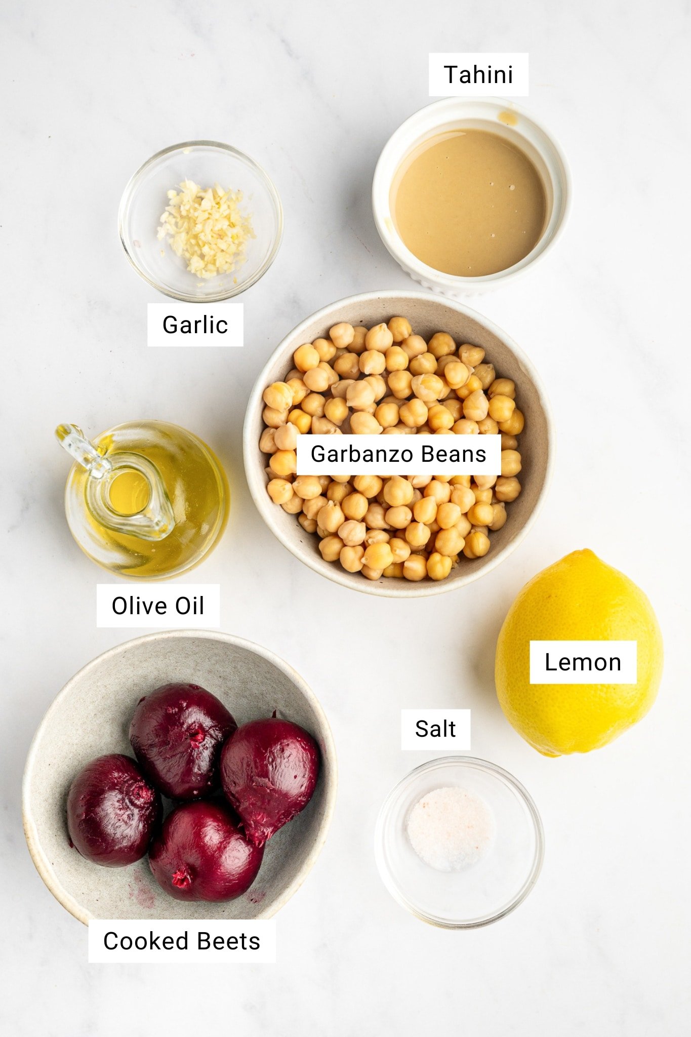 Labeled ingredients for beet hummus arranged on a light surface, including cooked beets, garbanzo beans, tahini, fresh garlic, lemon, olive oil, and salt.