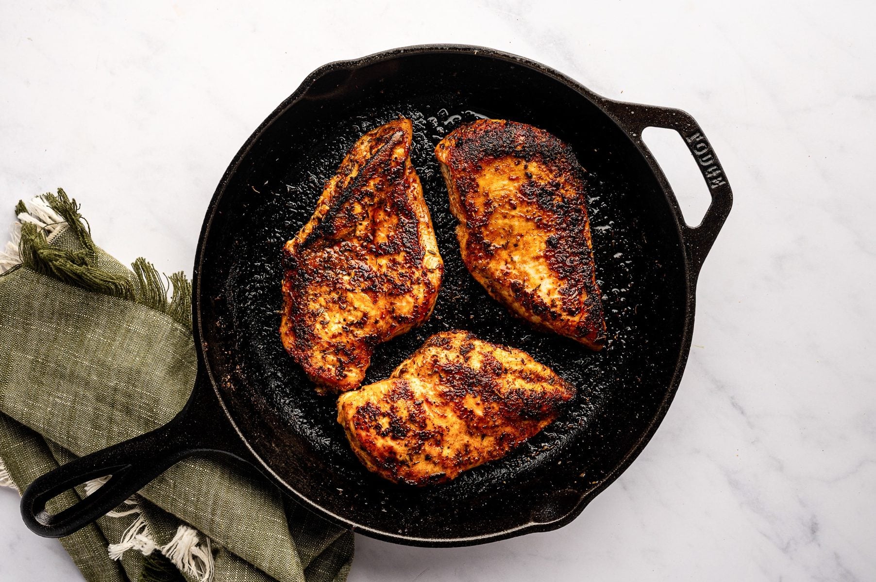 Overhead image of three cooked blackened chicken breasts in a black cast-iron skillet on a light gray marble surface. The chicken has a dark brown and blackened crust with caramelized seasoning patches and glossy juices in the pan. A muted olive green fringed kitchen towel sits below the skillet.