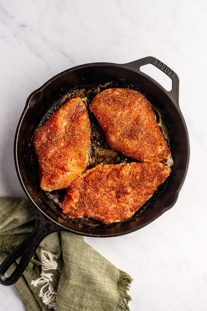 Overhead image of three seasoned raw chicken breasts in a black cast-iron skillet on a light gray marble surface. The chicken is coated in a red-orange blackening seasoning with visible dried herbs, and butter or oil is beginning to melt around the edges. A muted olive green fringed kitchen towel rests below the skillet.