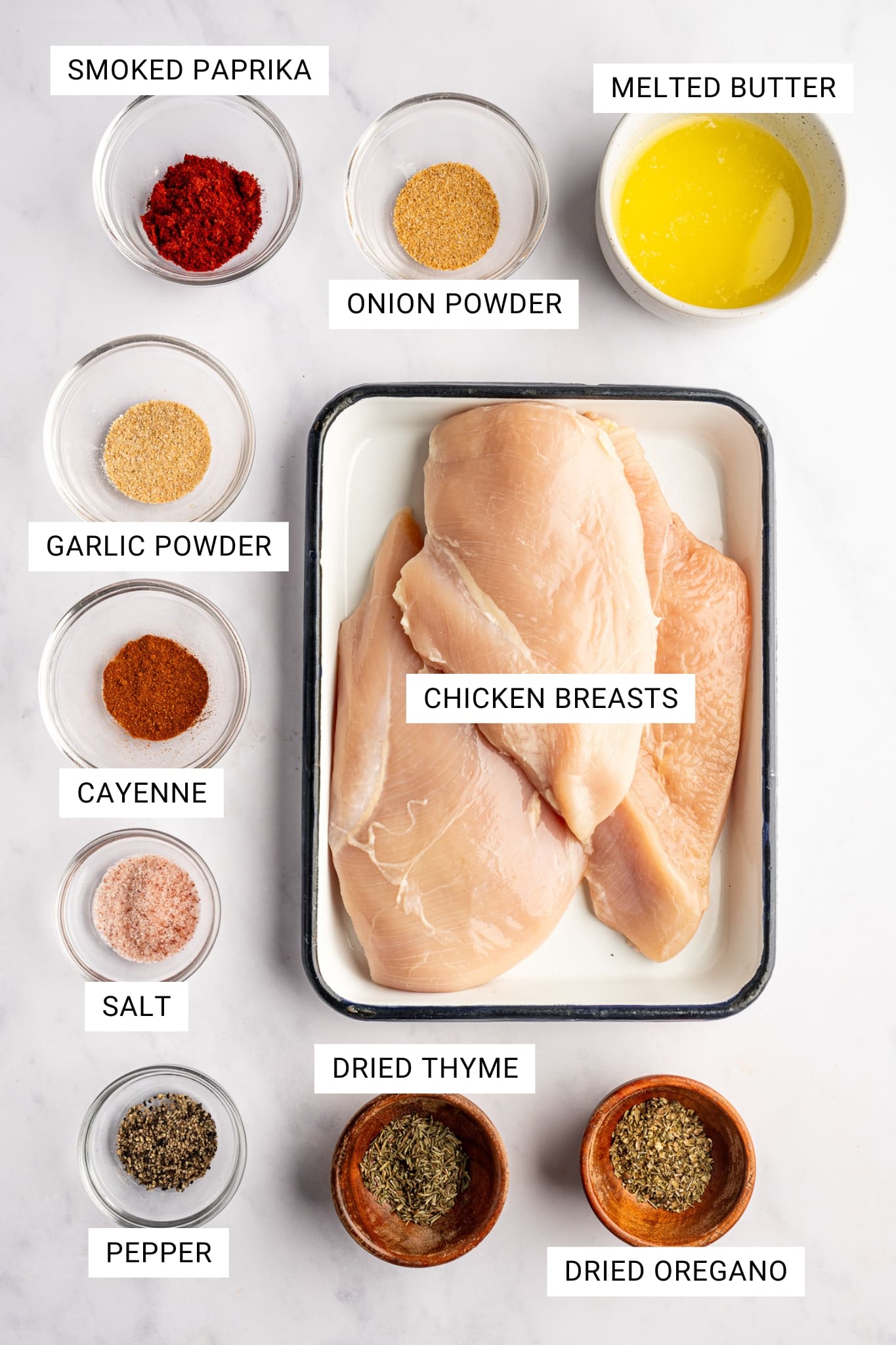 Overhead ingredients image for blackened chicken on a light gray marble surface. Raw chicken breasts sit in a white rectangular baking dish with a dark rim, surrounded by small clear glass bowls and small wooden bowls filled with smoked paprika, onion powder, garlic powder, cayenne, salt, pepper, dried thyme, dried oregano, and a white bowl of melted butter. White text labels identify each ingredient, including SMOKED PAPRIKA, ONION POWDER, MELTED BUTTER, GARLIC POWDER, CAYENNE, SALT, PEPPER, DRIED THYME, DRIED OREGANO, and CHICKEN BREASTS.