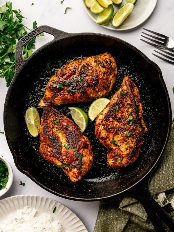 Overhead image of three blackened chicken breasts in a black cast-iron skillet garnished with chopped parsley and lime wedges. The skillet sits on a light gray marble surface with fresh parsley, a small white bowl of chopped herbs, a white plate of lime wedges, two silver forks, and a beige plate of white rice partially visible around it. A muted olive green towel is draped under the skillet.