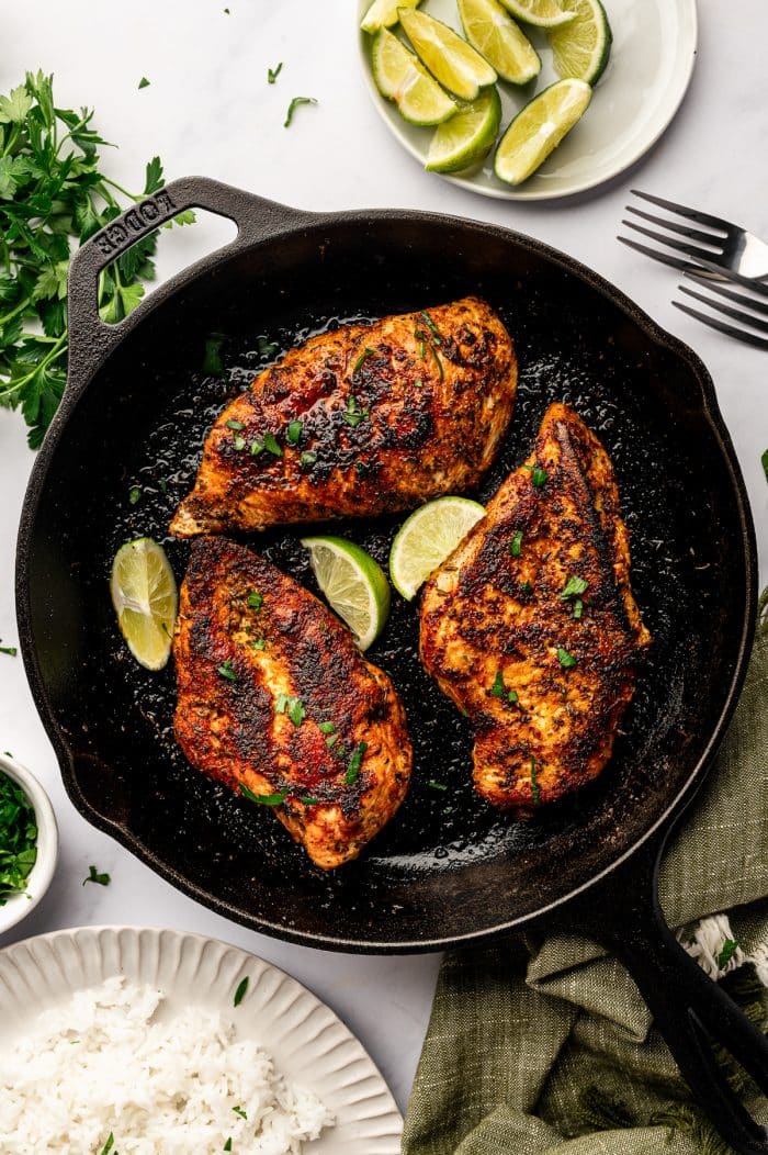 Overhead image of three blackened chicken breasts in a black cast-iron skillet garnished with chopped parsley and lime wedges. The skillet sits on a light gray marble surface with fresh parsley, a small white bowl of chopped herbs, a white plate of lime wedges, two silver forks, and a beige plate of white rice partially visible around it. A muted olive green towel is draped under the skillet.