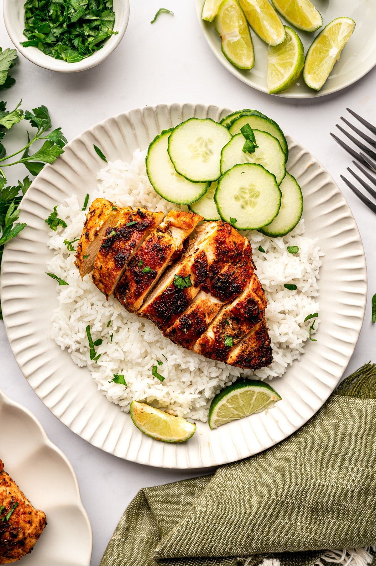 Overhead image of sliced blackened chicken served over white rice on a round beige plate with a fluted rim. Thin cucumber slices are arranged at the top of the plate, and lime wedges sit around the rice. Chopped parsley is sprinkled over the chicken and rice. Around the plate are a small white bowl of chopped herbs, a white plate of lime wedges, fresh parsley, two silver forks, and a muted olive green kitchen towel on a light gray marble surface.