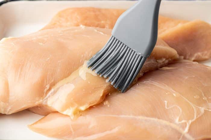 Close-up image of raw boneless skinless chicken breasts on a white surface while a gray silicone pastry brush brushes melted butter over the top. The chicken is pale pink and glossy from the butter.