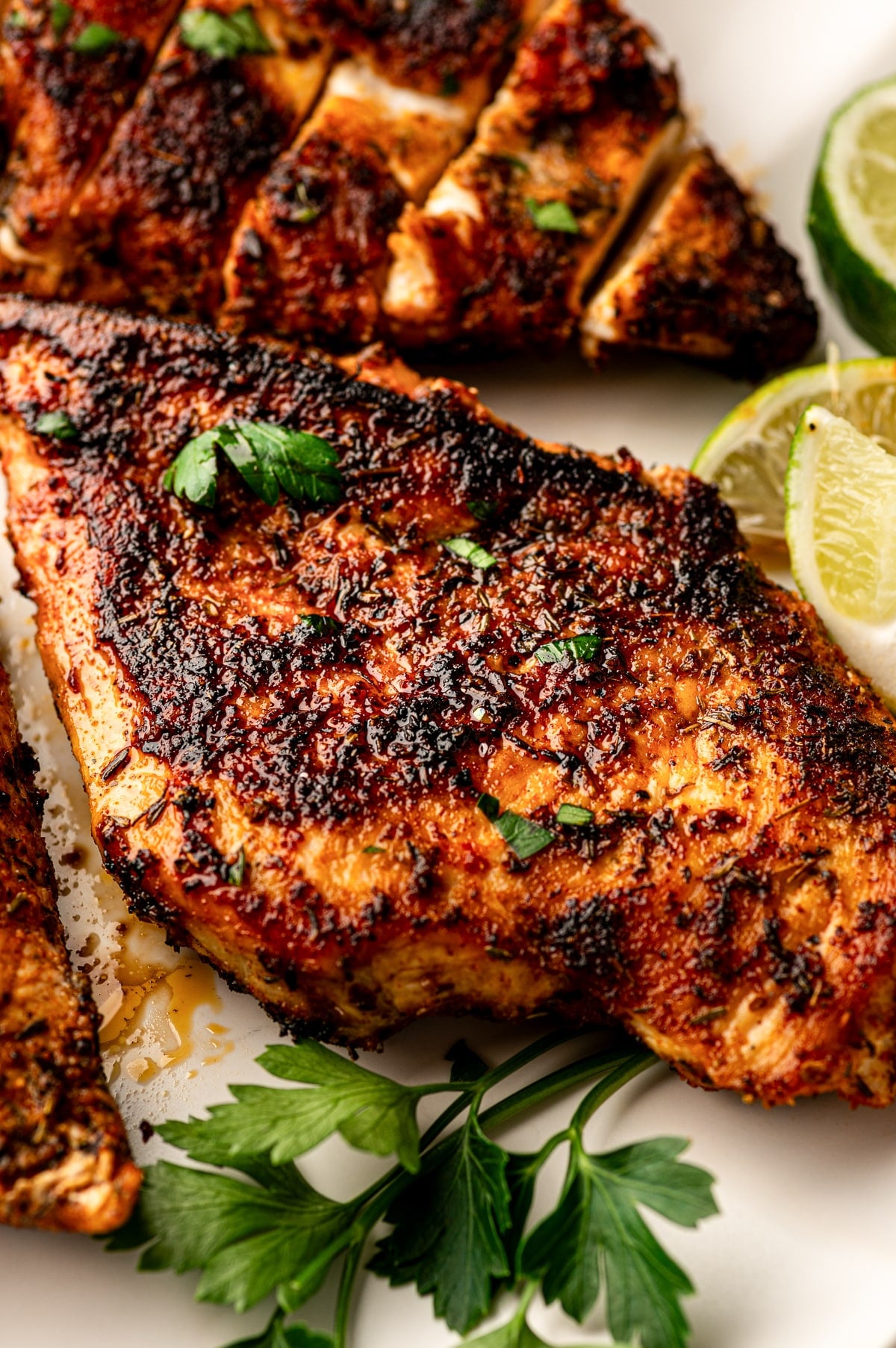 Close-up image of blackened chicken on a light plate with a dark, crusty exterior and glossy surface. One piece is sliced in the background, and lime wedges and fresh parsley are arranged around the chicken. The seasoning is deeply browned with visible herbs.