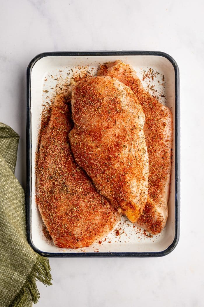 Overhead image of raw chicken breasts coated in blackened seasoning in a white rectangular baking dish with a dark rim. The seasoning is red and speckled with dried herbs. The dish sits on a light gray marble surface with a muted olive green fringed kitchen towel along the lower left side.