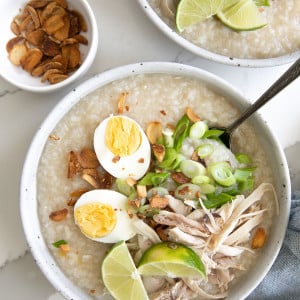 Bowls filled with cooked arroz caldo and garnished with fried garlic, scallions, hard-boiled egg, and shredded chicken.