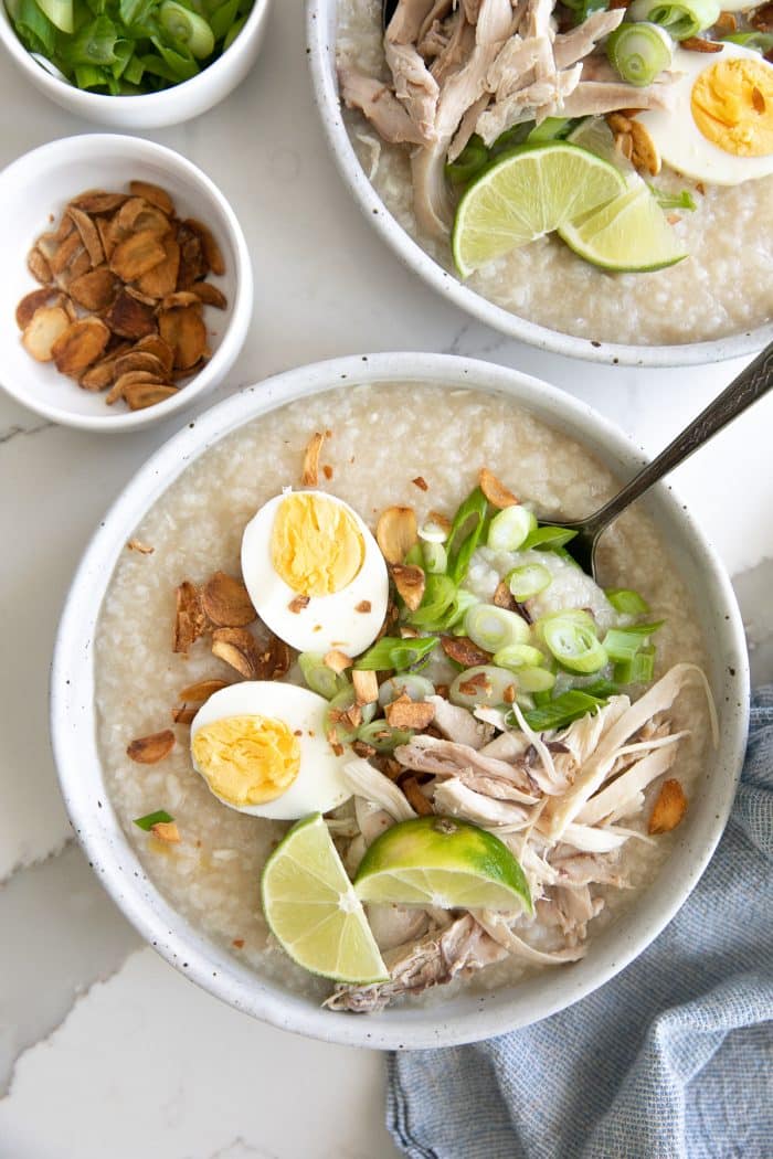 Bowls filled with cooked arroz caldo and garnished with fried garlic, scallions, hard-boiled egg, and shredded chicken.