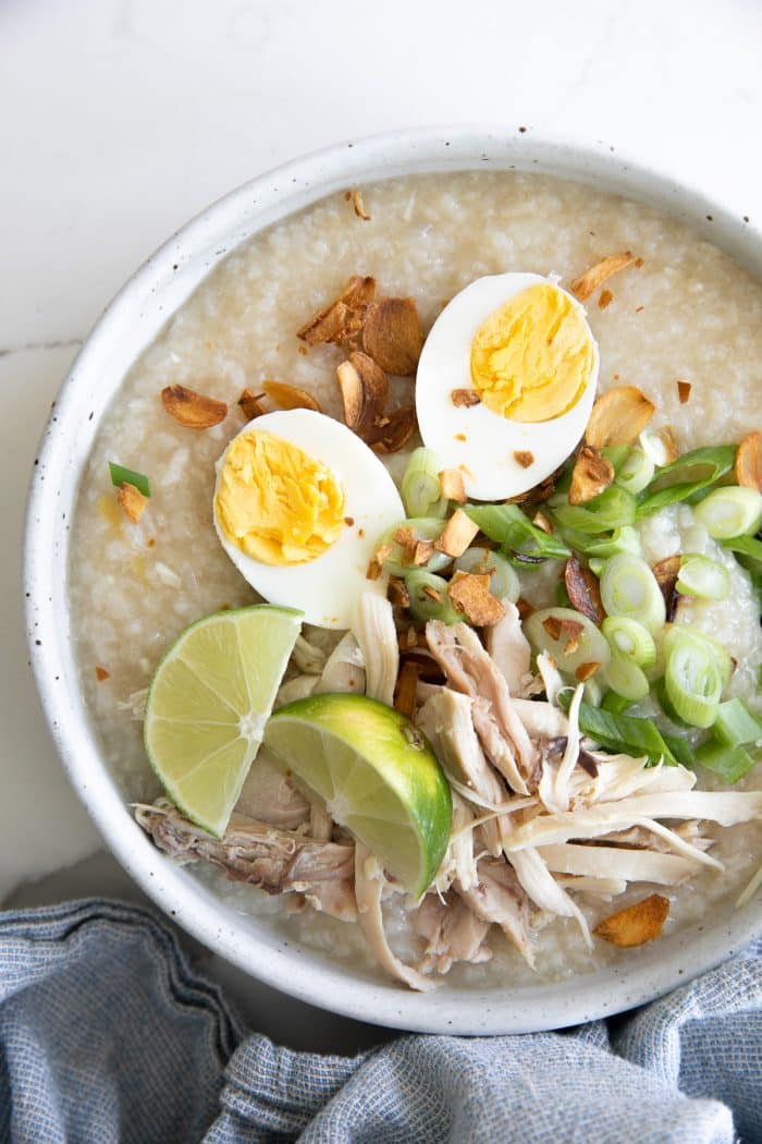 Bowls filled with cooked arroz caldo and garnished with fried garlic, scallions, hard-boiled egg, and shredded chicken.