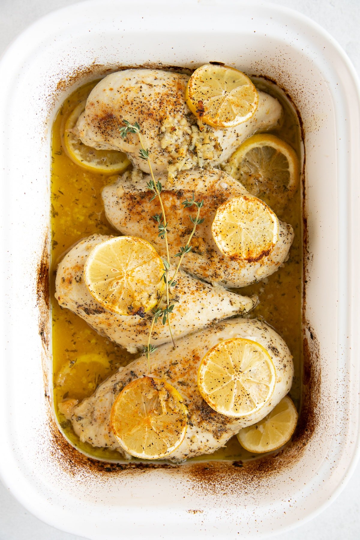 Four large chicken breasts in a white baking dish cooked in a buttery lemon, garlic, and herb sauce.