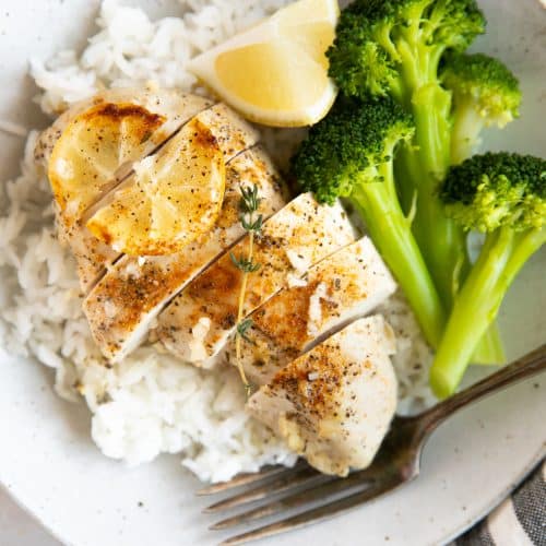 Juicy baked lemon chicken breast served on top of white rice with a side of cooked broccoli.
