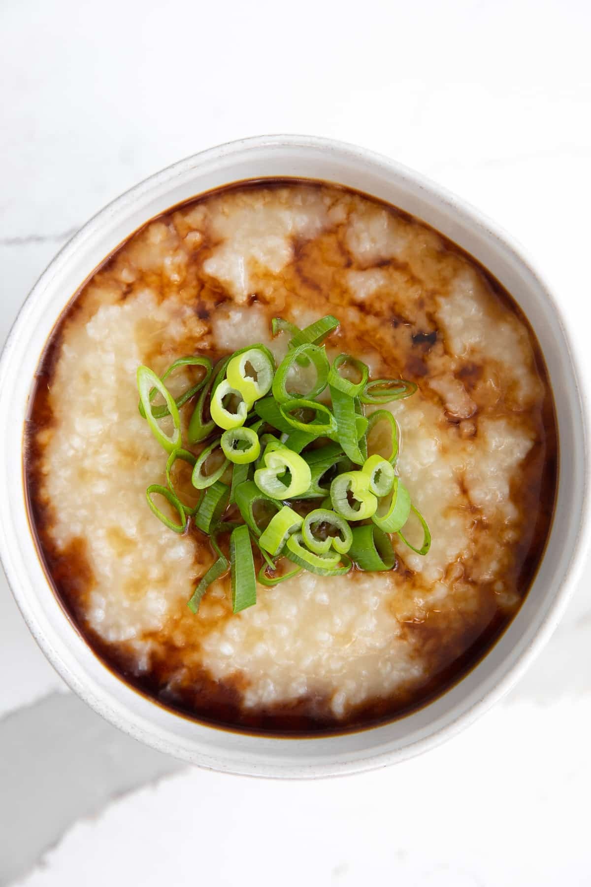 Small white bowl filled with Chinese rice porridge and garnished with soy sauce, sesame oil, and chopped green onions.