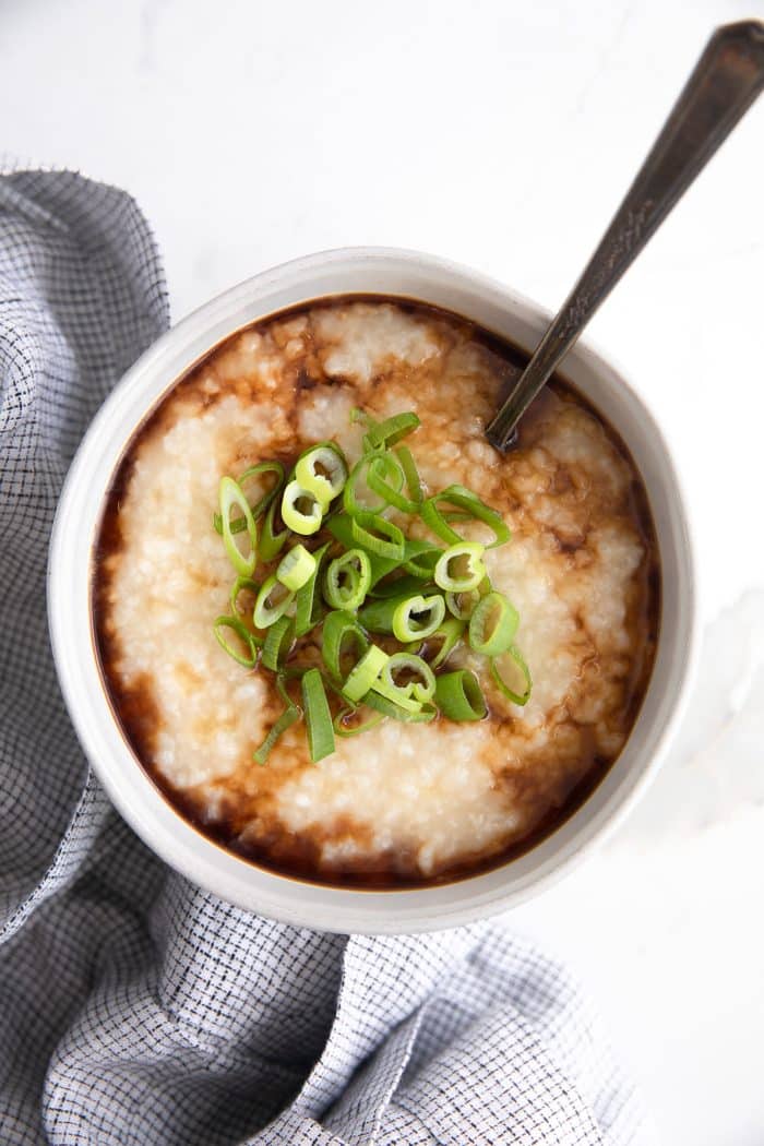 Simple homemade congee in a small white serving bowl topped with green onions and garnished and drizzled with sesame oil and soy sauce.