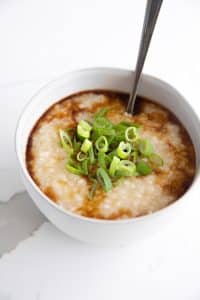 Simple homemade congee in a small white serving bowl topped with green onions and garnished and drizzled with sesame oil and soy sauce.