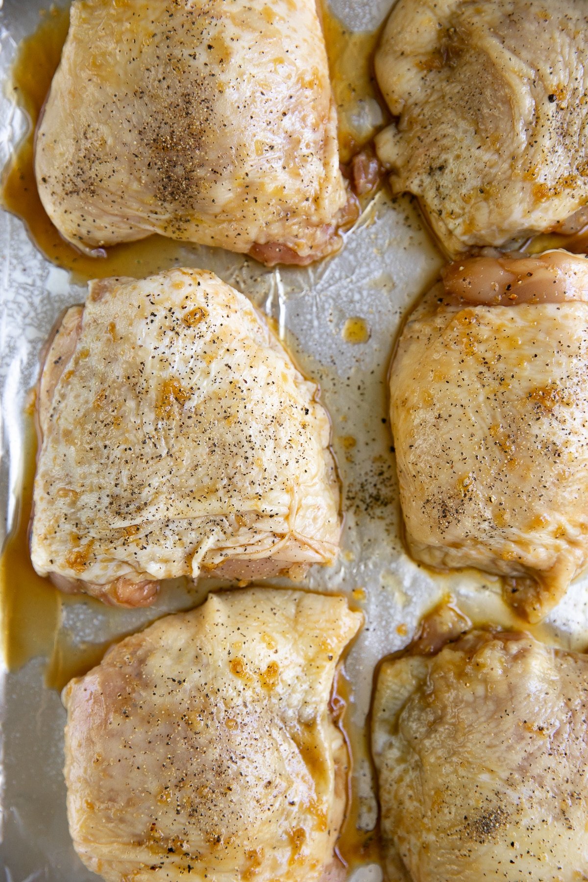 Six large skinned chicken thighs marinated in a honey soy sauce set on a large rimmed baking sheet lined with aluminum foil.